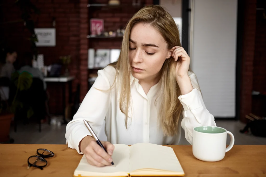 candid-shot-attractive-blonde-student-girl-white-blouse-doing-homework-workplace-home-writing-down-open-copybook-drinking-tea-having-serious-concentrated-facial-expression