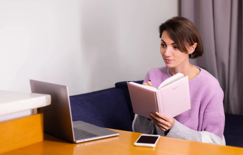 woman reading book and writing notes on laptop