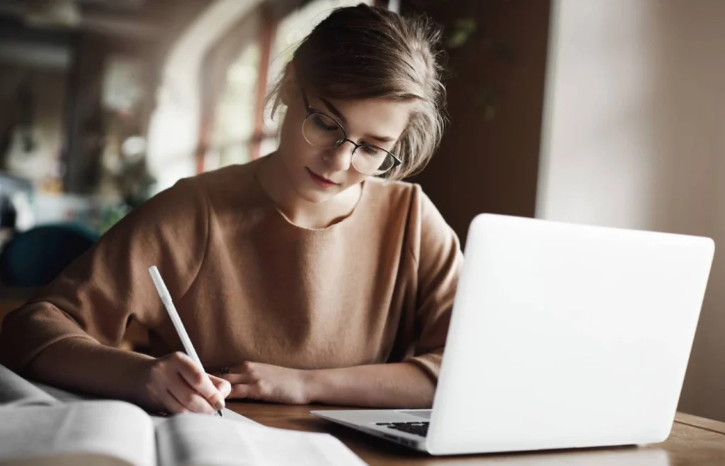 girl writing in notebook using laptop at desk